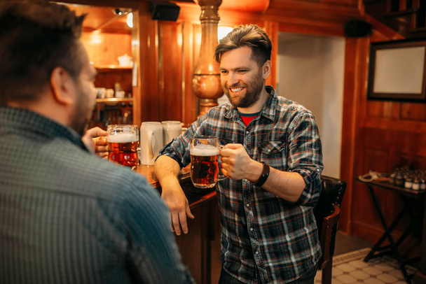 Two smiling male friends drinks beer at the counter in pub. Bearded men with mugs having fun in bar, good friends - Fotografie, Obrázek