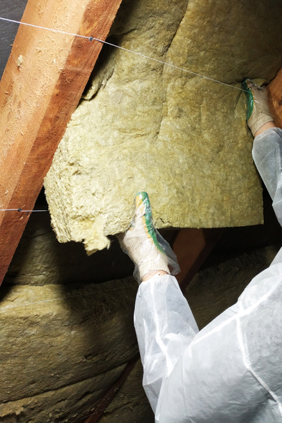 A man in a protective overalls puts mineral wool between the beam of the roof of the house for his warming from the cold - Photo, Image