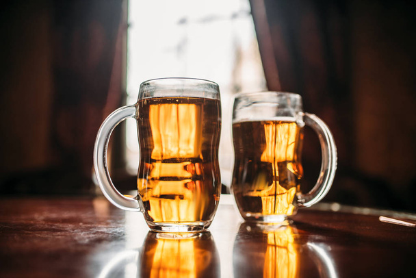Two beer mugs on wooden bar counter, macro view, nobody. Octoberfest symbol. Glasses with golden beverage on the table in pub, closeup, window with sunlight on background - Photo, Image