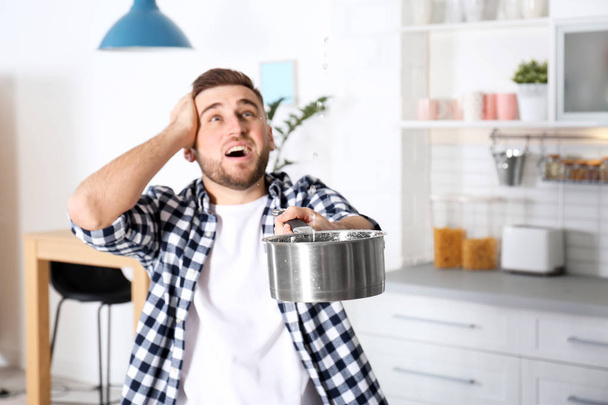 Emotional young man holding saucepan under water leakage from ceiling in kitchen, space for text. Plumber service - Φωτογραφία, εικόνα