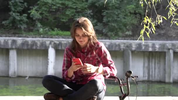 Subject ecological bicycle transport. Young Caucasian woman in shirt student sits resting in a park near the lake for rent orange bike uses a mobile phone autumn break in sunny weather technology - Footage, Video