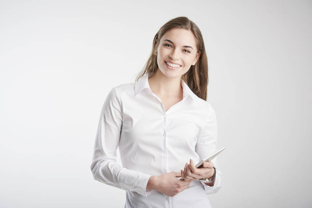 Studio shot of young businesswoman with toothy smile holding digital tablet in her hand while standing at isolated white background.  - Fotografie, Obrázek