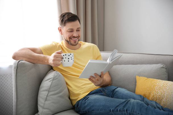 Handsome man with cup of coffee reading book on sofa at home - Foto, Bild