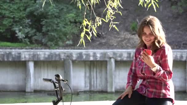 Temat ekologicznych rowerów transportu. Młoda kobieta kaukaski koszula student siedzi, odpoczynek w parku w pobliżu jeziora do wynajęcia pomarańczowy rower używa telefonu komórkowego Jesienna przerwa w technologii słonecznej pogody - Materiał filmowy, wideo