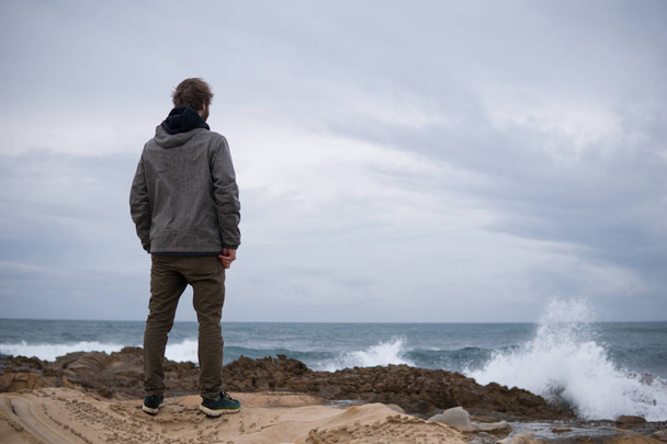 Ταξιδευτής στα βράχια κοντά στη θάλασσα κοιτάζοντας μακριά στον ορίζοντα. Ακτή βραχώδης Ατλαντικός ωκεανός και θυελλώδης καιρός. Όμορφος νέος Καυκάσιος τουρίστας άνθρωπος σε casual ρούχα σε εξωτερικούς χώρους στη φύση - Φωτογραφία, εικόνα