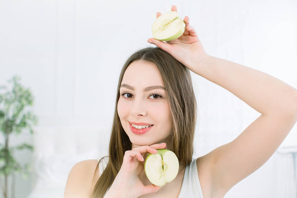 portrait of a young smiling beautiful girl with two halves of apples in her hands, above and below the face. Pretty Caucasian girl holding the halfs of apple and posing on the light background - Foto, immagini