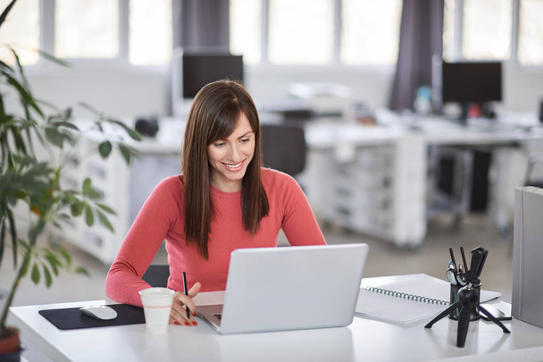 Beautiful smiling Caucasian businesswoman sitting in modern office and using laptop. - Photo, image