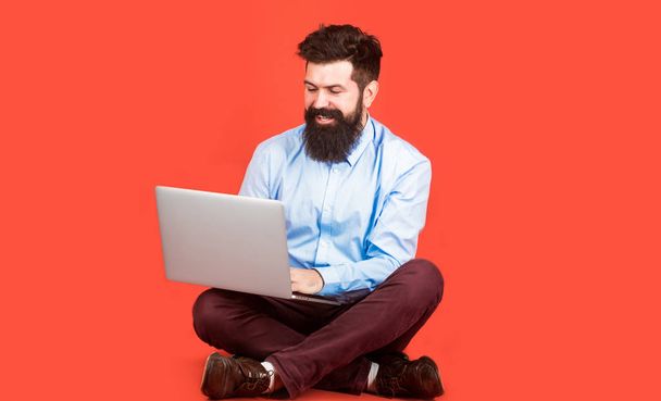 Mutlu genç ile katta oturan ve kırmızı zemin üzerine dizüstü bilgisayar kullanan adam. Dizüstü bilgisayar tutarak. Genç işadamı, onun dizüstü bilgisayar kullanarak pc. Yakışıklı gülümseyen erkek çalışan lapt sakallı - Fotoğraf, Görsel