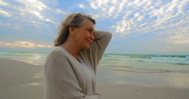 Vooraanzicht van een actieve Kaukasische bejaarde vrouw glimlachend en permanent op het strand terwijl de weg op zoek en het passeren van haar hand door haar haren 4k - Video