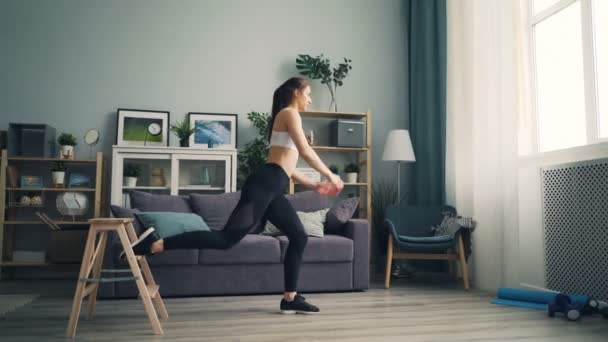 Attractive girl squatting on 1 leg doing sports indoors at home using furniture - Кадры, видео