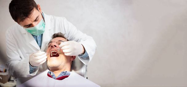 Young male dentist holding a tooth mirror and dental pick, performing dental exam on a mature male patient in dental clinic. Wide ratio image with copy space on the right side - Фото, зображення