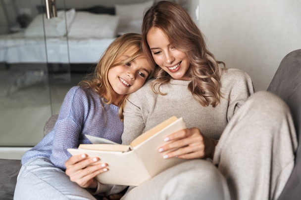 Young woman with her little sister indoors at home reading book. - Fotoğraf, Görsel