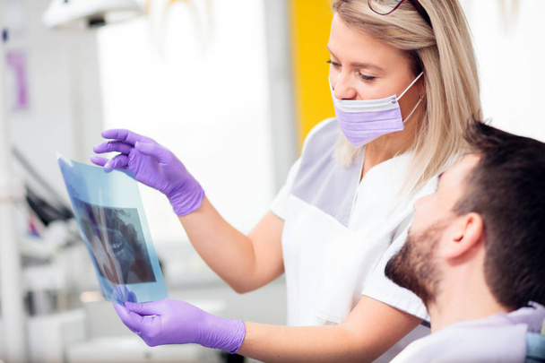 Νεαρή γυναίκα οδοντίατρος δείχνει μια εικόνα ακτινογραφία σε έναν αρσενικό ασθενή μετά από μια επιτυχημένη παρέμβαση - Φωτογραφία, εικόνα