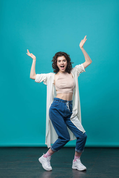  beautiful young brunette woman in casual style screaming with raised hands and posing against blue wall background  - Photo, Image