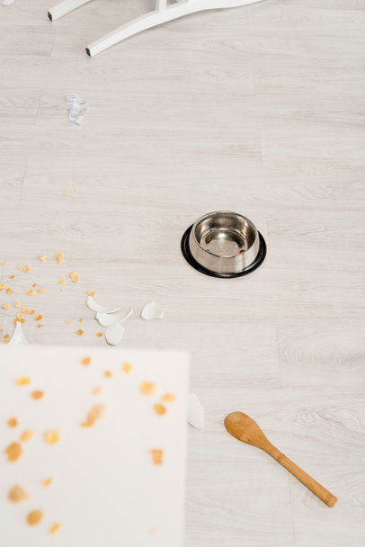 selective focus of metal bowl, wooden spoon and broken dishes on floor in kitchen  - Foto, afbeelding