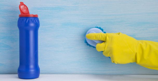 mão em uma luva amarela escovando a superfície, de pé ao lado de um frasco azul com agente de limpeza, em um fundo leve
 - Foto, Imagem