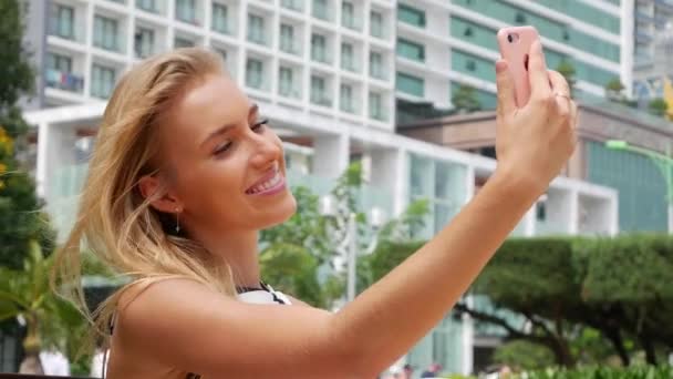 Beautiful slim woman with long blonde hair in black and white dress making selfie on mobile phone over background the park. - Záběry, video