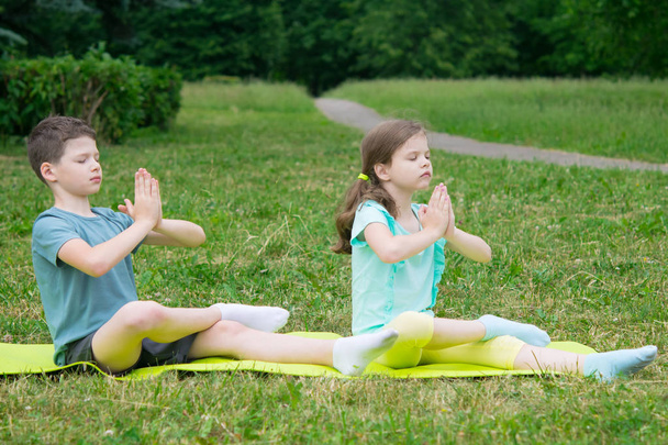 boy and girl do yoga, eyes closed, in the open air, on a background of green grass, sitting on a gymnastic mat - Fotografie, Obrázek