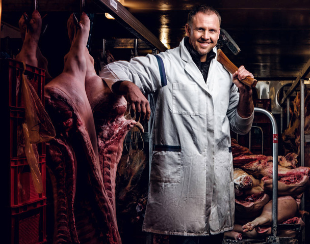 Boucher en chemise de travail blanche tenant la hache debout dans un entrepôt frigorifique au milieu de carcasses de viande
 - Photo, image