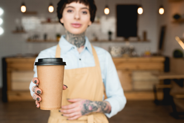 selective focus of attractive waitress in apron holding paper cup and looking at camera - Foto, imagen