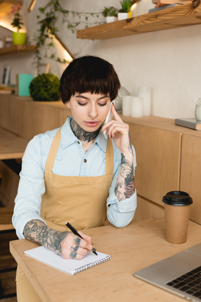 concentrated waitress talking on smartphone and writing in notebook - Photo, Image