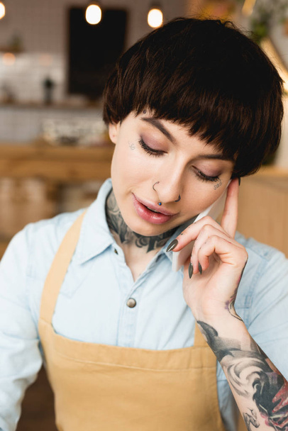 ελκυστικές τατουάζ σερβιτόρα μιλώντας στο smartphone και κοιτάζοντας προς τα κάτω - Φωτογραφία, εικόνα