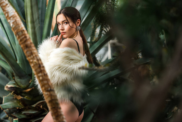 Sexy young woman in faux fur coat and underwear near plants in botanical garden - Фото, изображение