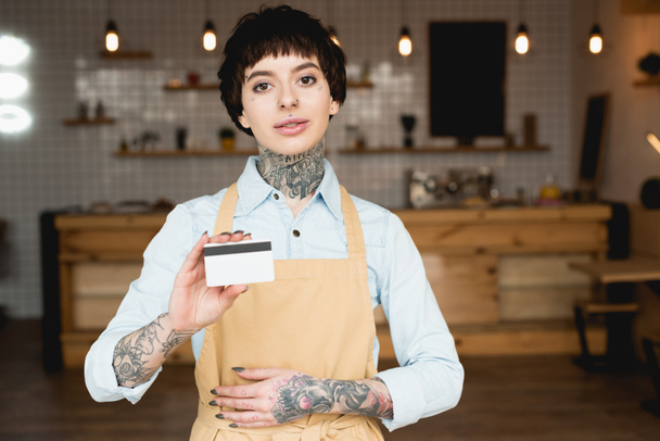 atractiva camarera en delantal con tarjeta de crédito y mirando a la cámara
 - Foto, imagen