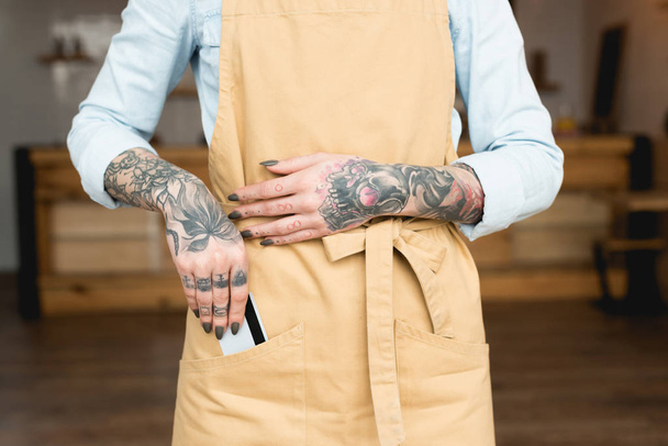 vista parcial de camarera tatuada poniendo tarjeta de crédito en bolsillo delantal
 - Foto, Imagen
