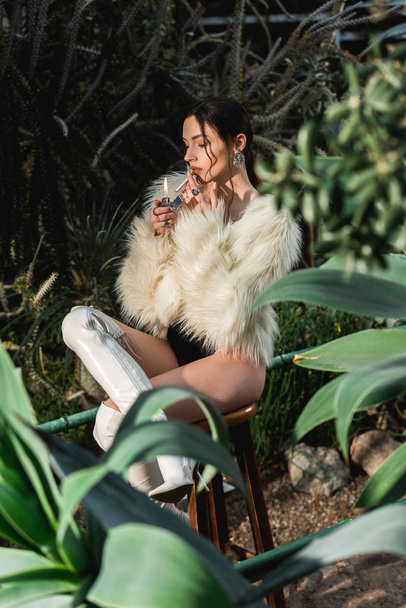 Sexy joven mujer en piel sintética abrigo iluminación cigarrillo en el jardín botánico
 - Foto, Imagen
