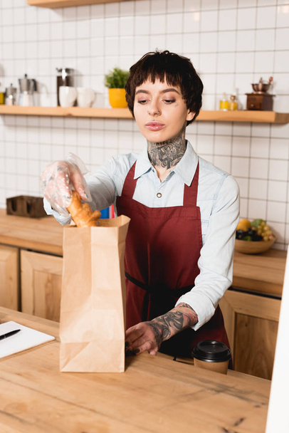 pretty barista in apron putting croissant into paper bag while standing at bar counter - Photo, Image