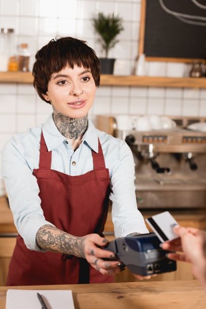 smiling cashier holding payment terminal near buyer with credit card - Photo, Image