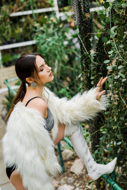 Confident sexy girl in faux fur coat touching green foliage in orangery - Фото, изображение