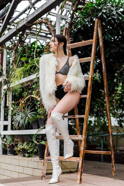 Sexy peinzende meisje in faux fur jas en ondergoed staande in de buurt van houten ladder in de Oranjerie - Foto, afbeelding