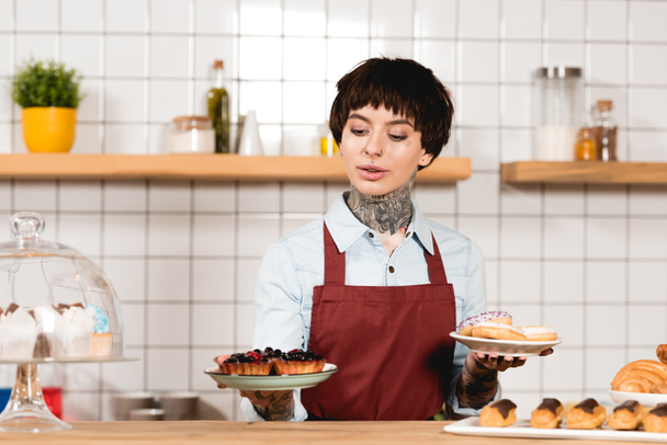 atraente barista segurando pratos com deliciosa pastelaria no café
 - Foto, Imagem