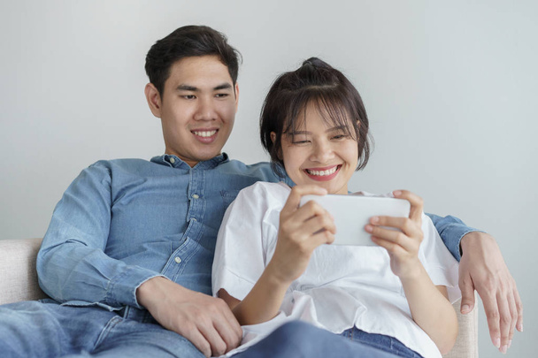 Happy young love asian couple sitting on a couch at home, looking at mobile phone - Foto, Imagem