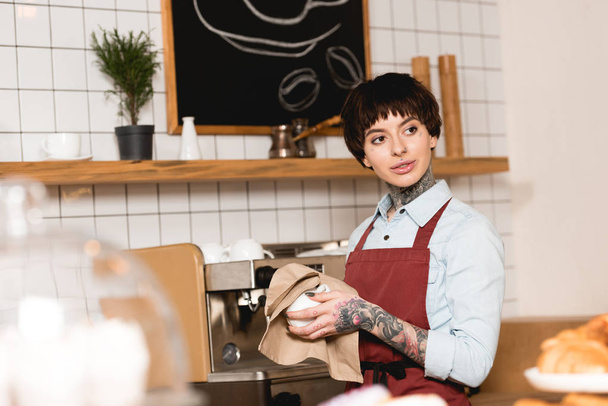 selective focus of pretty barista wiping cup while standing near espresso machine - Photo, Image