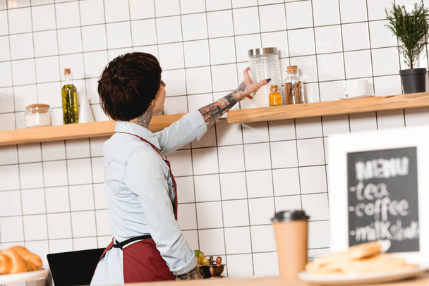 barista taking glass jar from wooden shelf in coffee shop - Фото, изображение