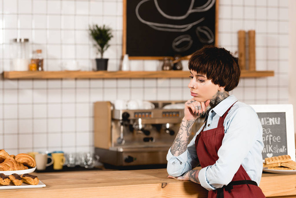 pensive barista in apron standing near wooden bar counter in coffee shop - Foto, Imagen