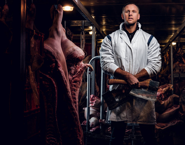 Macellaio in abiti da lavoro in posa con due assi in un magazzino refrigerato in mezzo alle carcasse di carne
 - Foto, immagini