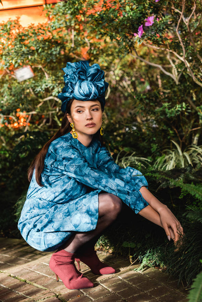 Traurige nachdenkliche Frau in blauem Kleid und Turban sitzt in Orangerie - Foto, Bild