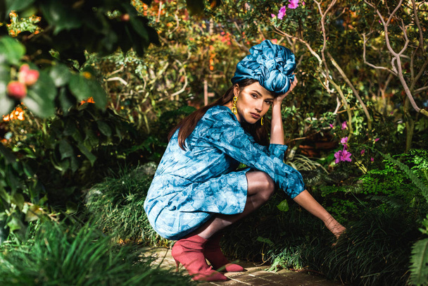 Triest peinzende vrouw in blauwe jurk en tulband zitten in de botanische tuin - Foto, afbeelding