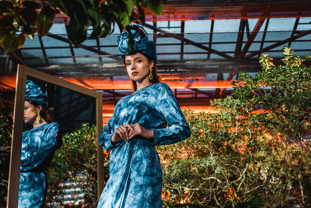 Beautiful woman in blue turban and dress standing near mirror in orangery - Foto, Bild