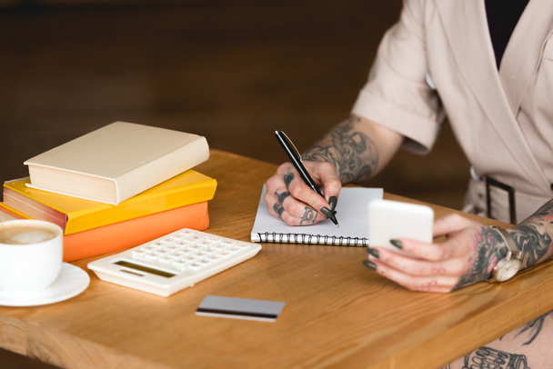 visão parcial de empresária tatuada escrevendo em notebook e segurando smartphone
 - Foto, Imagem