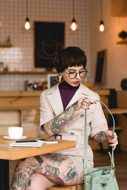 trendy, attractive businesswoman in eyeglasses holding bag while sitting in cafe - Photo, Image