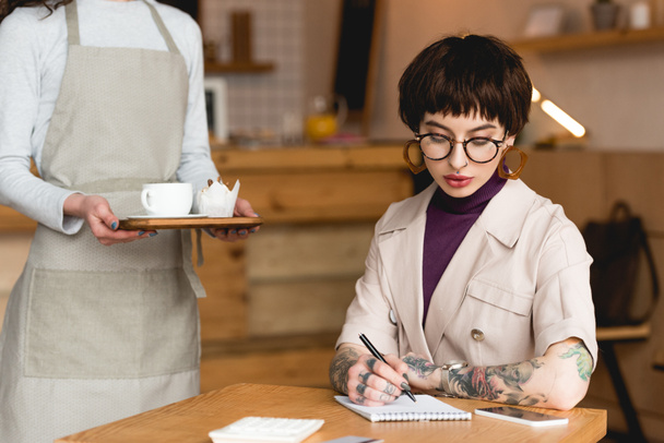 частичный взгляд на официантку, держащую поднос рядом с модной деловой женщиной, сидящей за столом и пишущей в блокноте
 - Фото, изображение