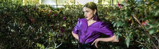 Panoramic shot of elegant young woman in purple blouse standing with arms akimbo in orangery - Fotoğraf, Görsel