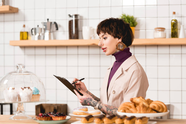 mooie zakenvrouw schrijven in klembord terwijl staande op toog in coffeeshop - Foto, afbeelding
