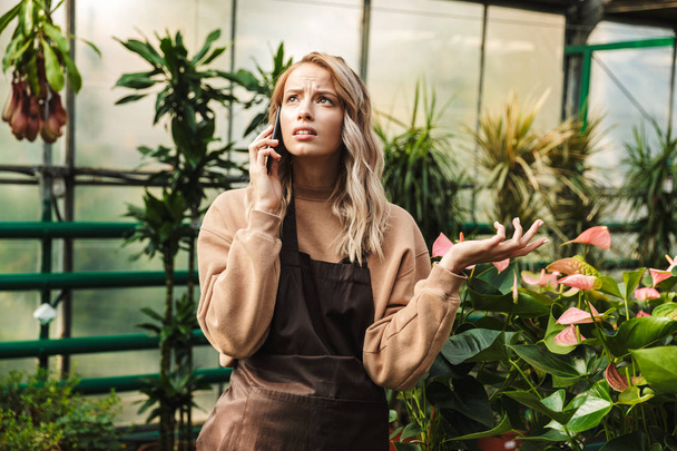 Beautiful gardener woman posing in the nature greenhouse garden talking by mobile phone. - 写真・画像