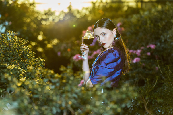 Impresionante joven en blusa púrpura sosteniendo copa de vino en jardín botánico
 - Foto, Imagen